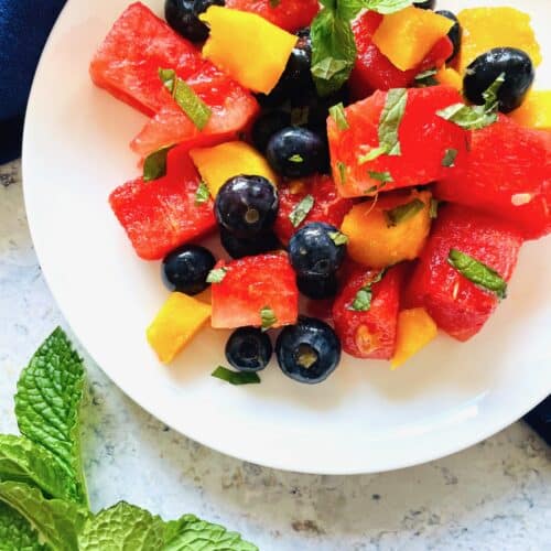 watermelon fruit salad with mango and blueberries recipe on a white plate with fresh mint leaves in the bottom left hand corner