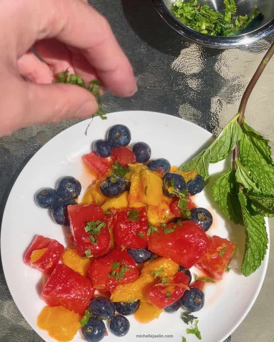 watermelon fruit salad with mango and blueberries on a plate being sprinkled with chopped fresh mint leaves
